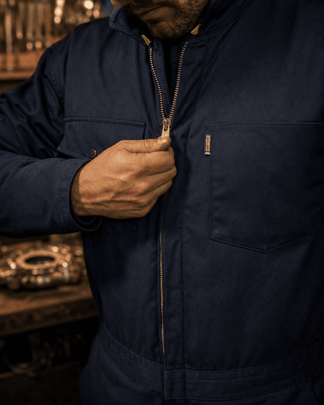 A close-up of a person wearing a dark navy blue work jacket, pulling the zipper with their hand. The jacket features a chest pocket with a small vertical metal brand tab and a zippered chest pocket.